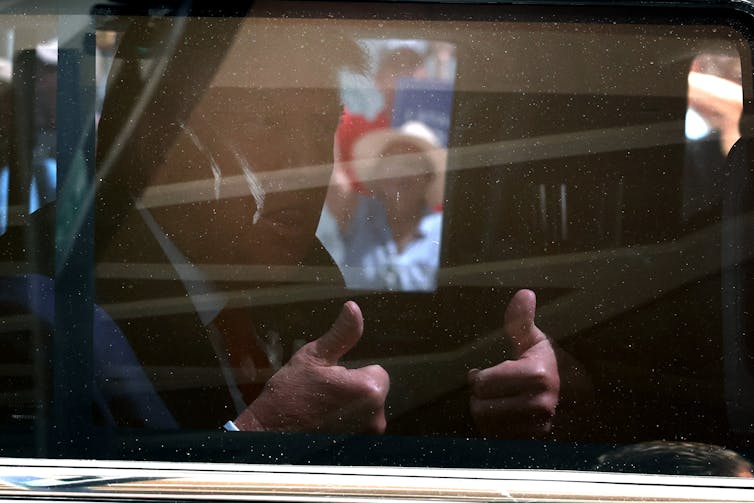 Former President Donald Trump is seen behind a glass window with two thumbs up. The photo is dark, showing the interior of a car.