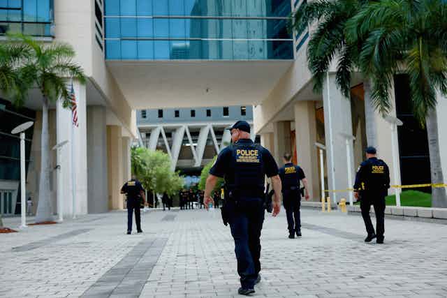 Four men wearing dark clothing with the yellow word police on it walk through an empty plaza that has palm trees, in front of a large, white building. 