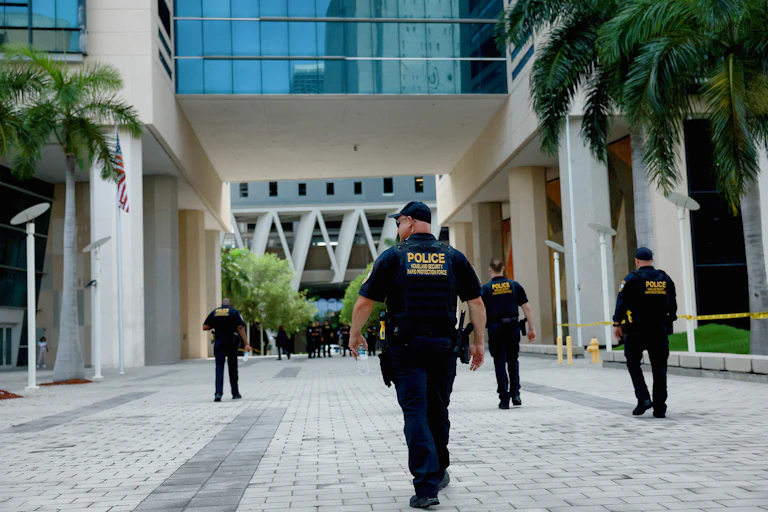 Four men wearing dark clothing with the yellow word police on it walk through an empty plaza that has palm trees, in front of a large, white building.