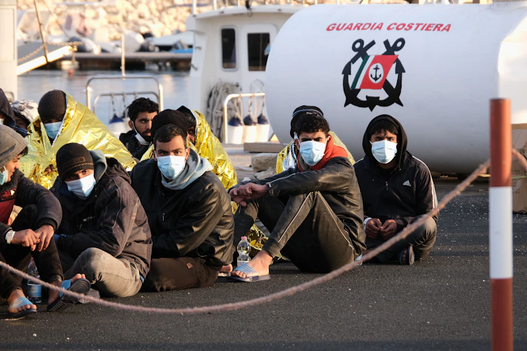 Migrantes sentados en el suelo con mascarilla miran a cámarra.