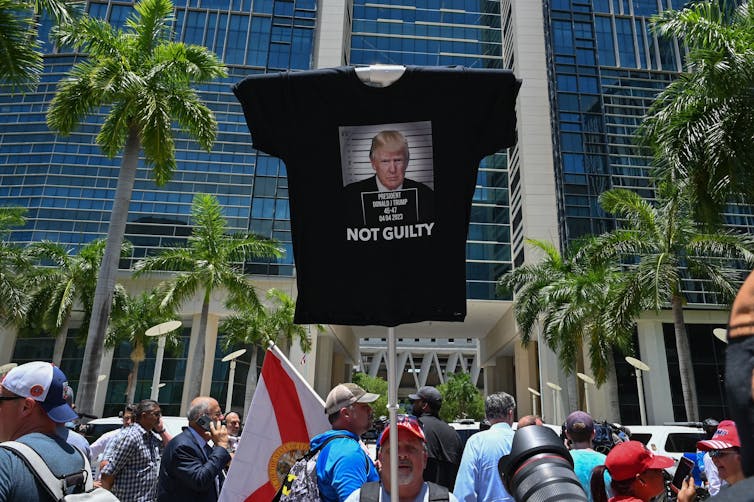 Un grupo de personas se aglomera fuera de un edificio con palmeras y uno sostiene una camiseta negra con la imagen de un hombre con un traje que dice 'no culpable'.