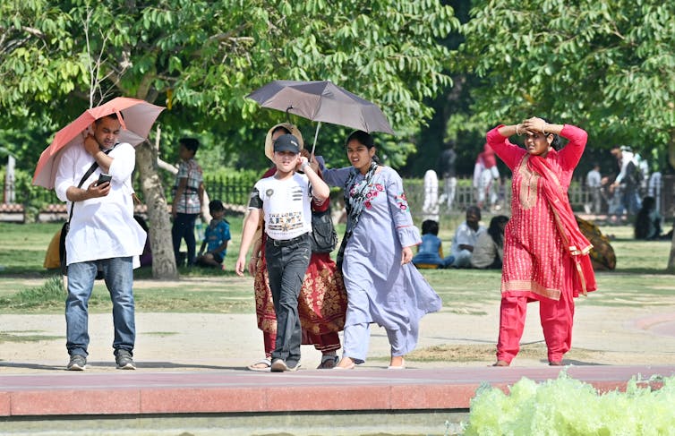 Трое взрослых ходят под зонтами, которые защищают их от солнца.  Женщина без зонта закрывает глаза руками в жаркий день, а мальчик носит шляпу.