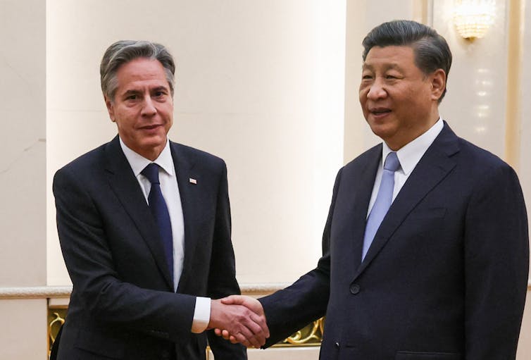 Two men, wearing suits and ties stand side-by-side and shake hands.
