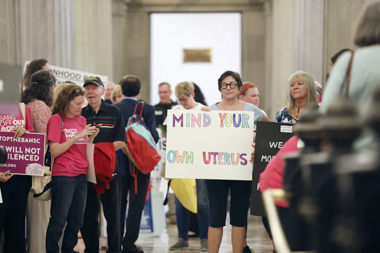 Protesters gather in a small crowd holding signs including