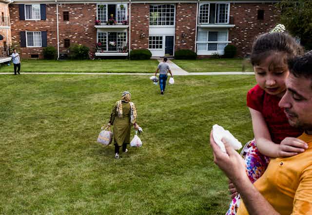 Two people carry groceries across a lawn towards an apartment complex. In the foreground a man holds a little girl.