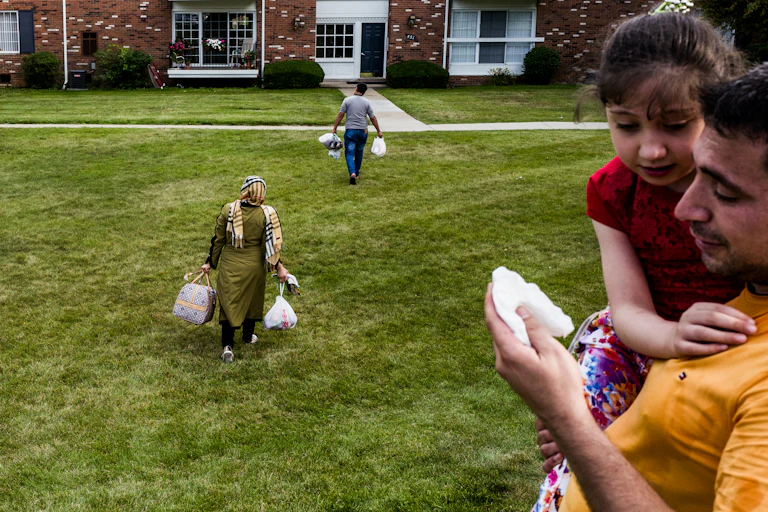 Two people carry groceries across a lawn towards an apartment complex. In the foreground a man holds a little girl.