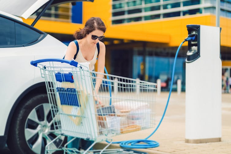 A woman unloads a shopping cart in a parking lot and puts items into her EV, which is charging from a public charger.