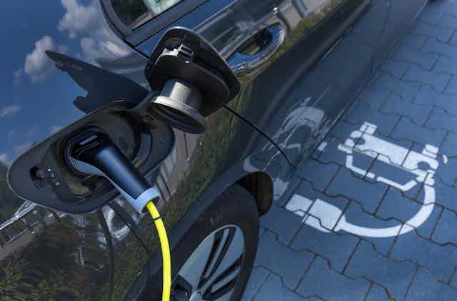 An electric car with yellow charging cord is parked in space marked with an EV charging symbol (a car with a plug). The sky is reflected in the vehicle's shiny side.