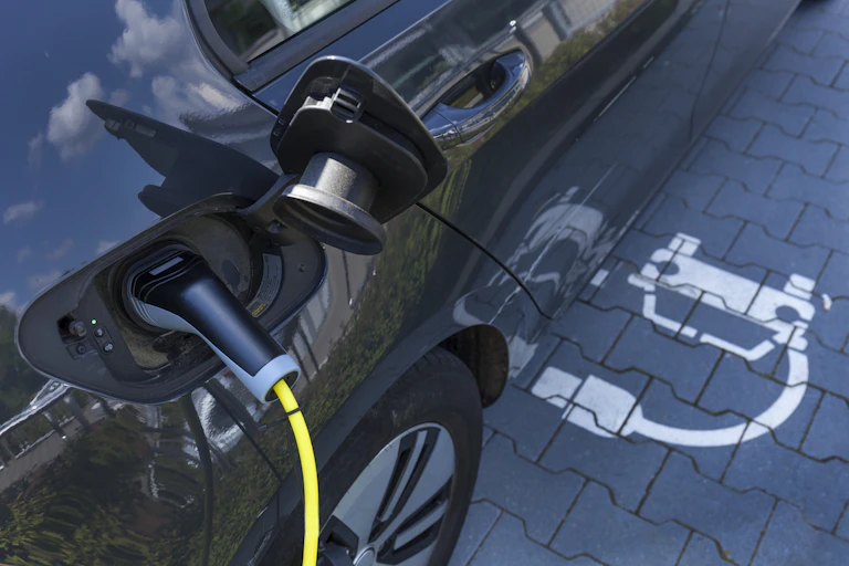 An electric car with yellow charging cord is parked in space marked with an EV charging symbol (a car with a plug). The sky is reflected in the vehicle's shiny side.