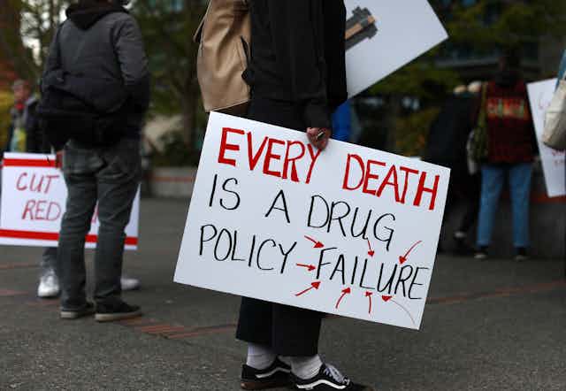 A person whose head is out of frame holds a sign reading 'Every death is a drug policy failure'