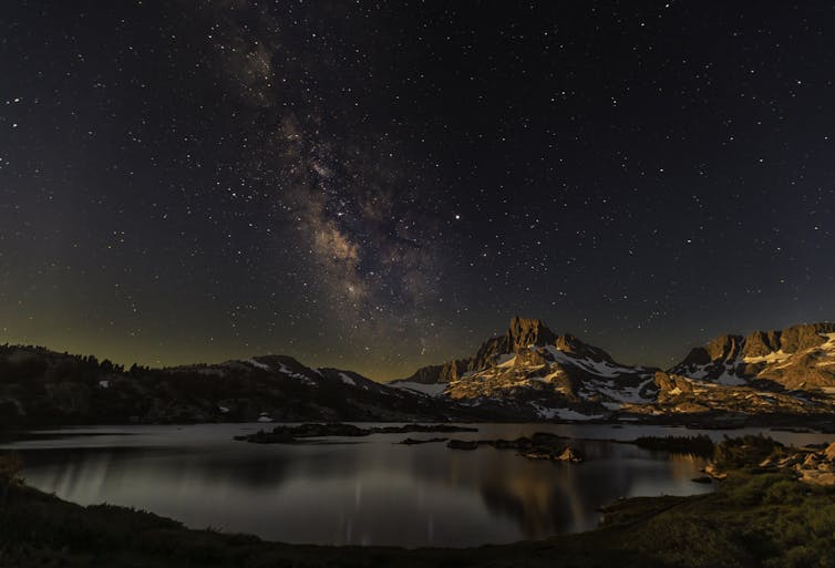 A lake under snow-speckled mountains, with lots of stars visible in the night sky.