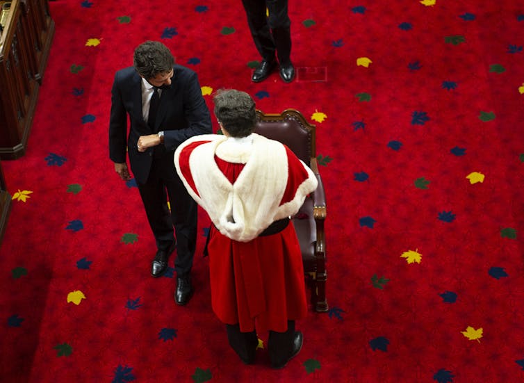A dark-haired man in a suit elbow bumps a man in a red robe lined with white fur against a crimson carpet.