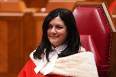 A woman with thick dark hair smiles as she sits in an ornate courtroom. She is in a red robe lined with white fur.