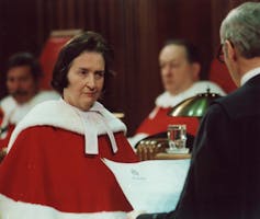 A woman with dark hair looks at a man reading to her. She wears a red robe lined with white fur.