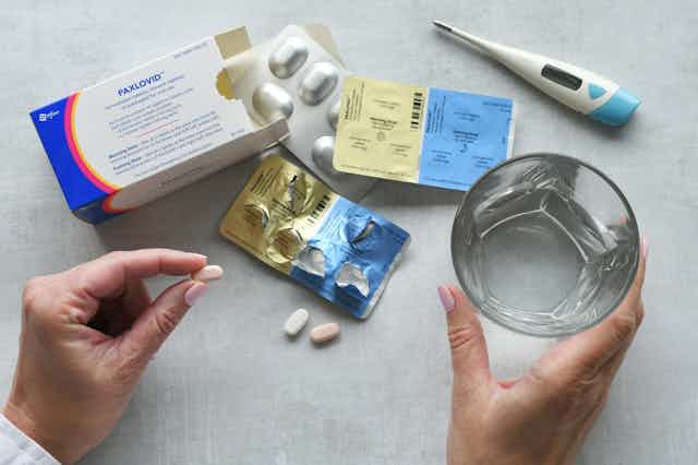 Woman taking Paxlovid, pill in one hand, glass of water in other, medicine and thermometer in background