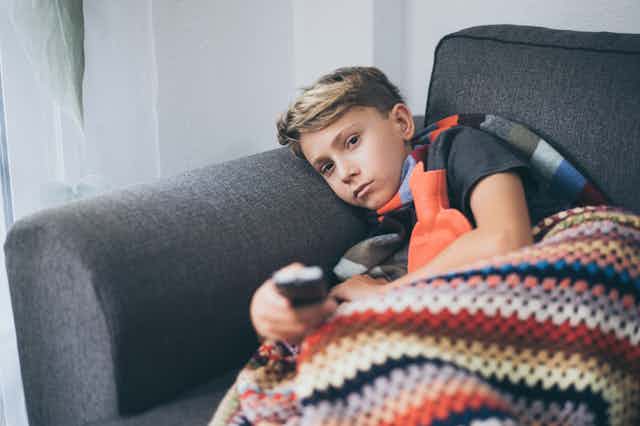 A sick boy lies on a couch with a blanket and tv remote.