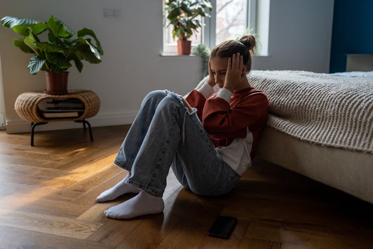 A teenage girl sits on the floor against her bed.