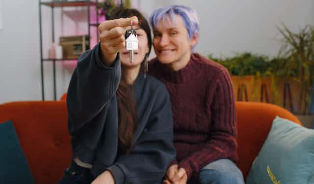 Two women sit on a couch, one holds a set of house keys towards the camera