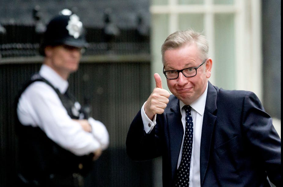 A white man wearing glasses and a suit and tie raises his thumb towards the camera, smirking.