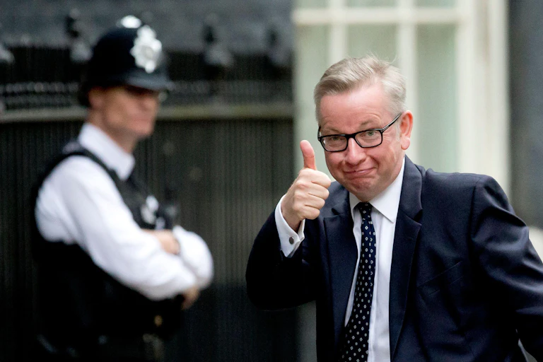 A white man wearing glasses and a suit and tie raises his thumb towards the camera, smirking.