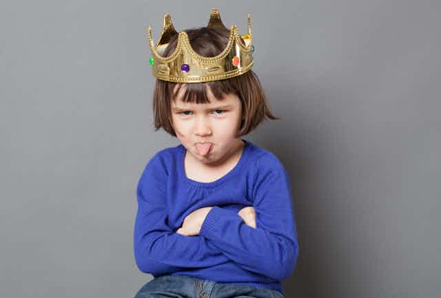 Cheeky preschool child with golden crown on head folding arms and sticking out tongue.