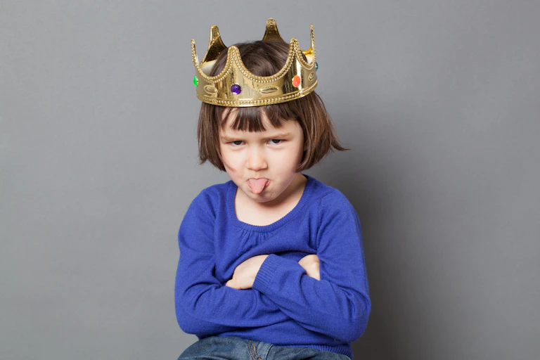 Cheeky preschool child with golden crown on head folding arms and sticking out tongue.