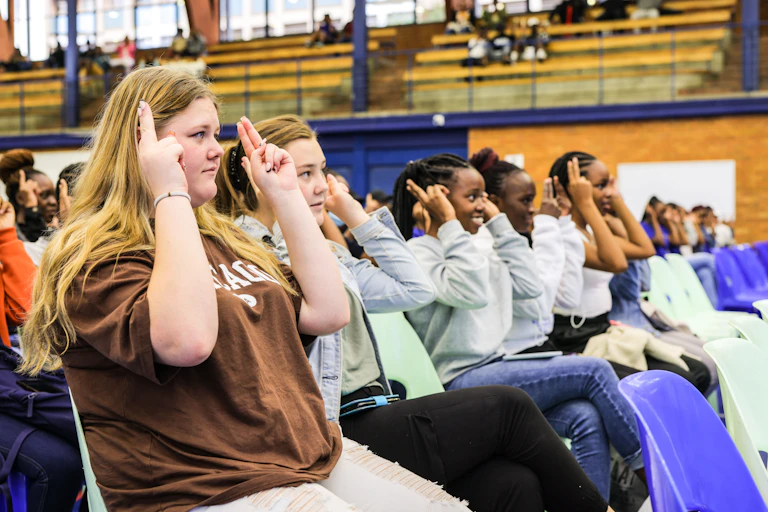 A row of young people in casual clothes are pictured holding two fingers on each hand up to their heads