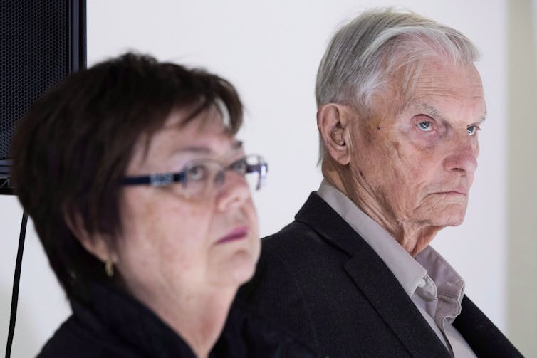 A grey-haired older man looks sombre as he stands next to a woman with short dark hair wearing glasses.