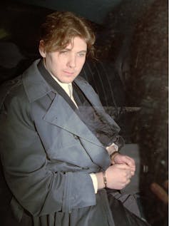 A man in handcuffs and a grey coat sits in the back of a police cruiser.