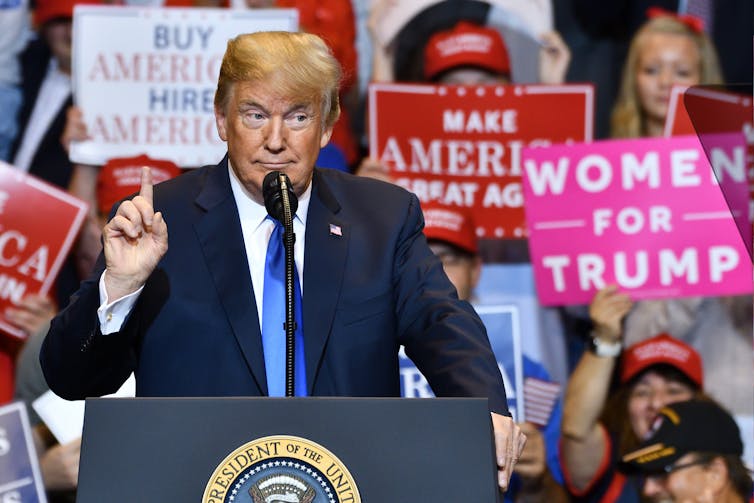 A man with orange face and hair stands behind a podium with a microphone raising his index finger with a doubtful expression.