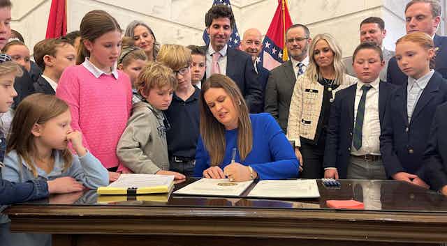 A woman with long brown hair wearing a longsleeved blue dress signs and official document while surrounded by children and adults, some wearing suits.