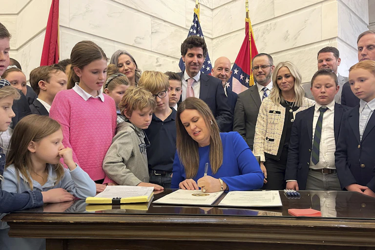 A woman with long brown hair wearing a longsleeved blue dress signs and official document while surrounded by children and adults, some wearing suits.