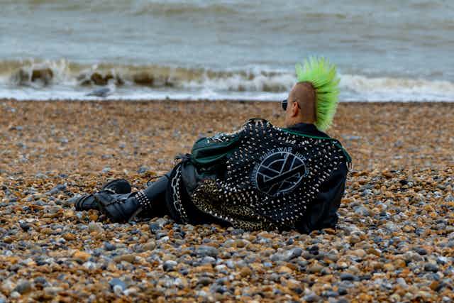 Punk allongé à la plage de galets. Il est coiffé d'une crête verte sur le sommet du crâne.
