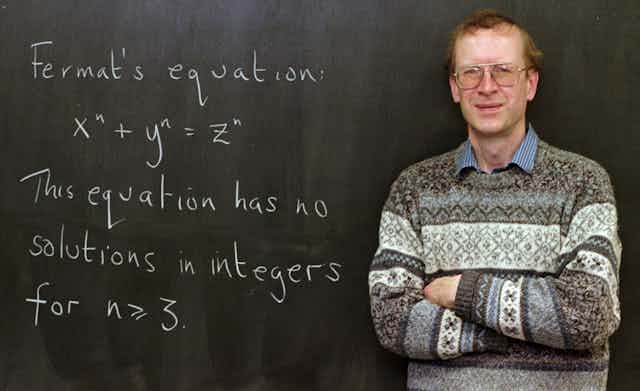 A man wearing a sweater and glasses stands with his arms crossed in front of a blackboard reading "Fermat's equation: x^n + y^n = z^n. This equation has no solutions in integers for n is greater than or equal to 3"