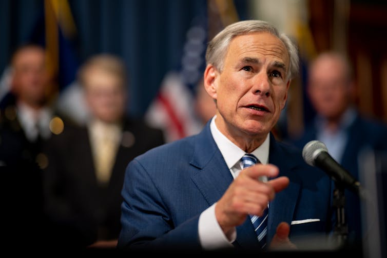 A gray-haired man in a blue suit and white shirt, talking while gesturing.