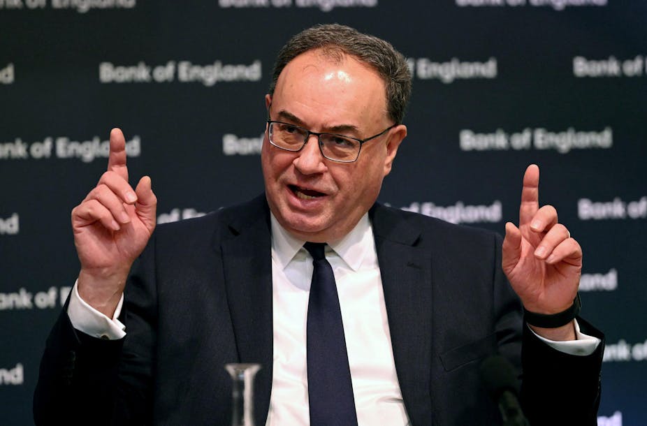 Man in dark suit, white shirt, glasses, hands up, fingers pointing upwards, black background with "Bank of England" in white letters.