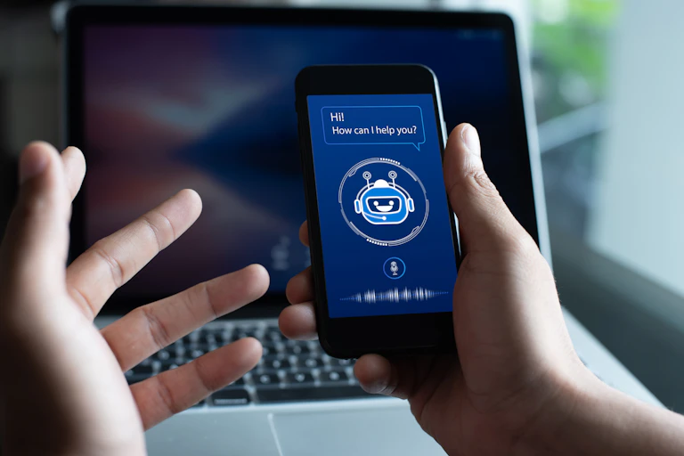 two hands holding a smartphone with the face of a robot on the screen and the text HI HOW CAN I HELP YOU