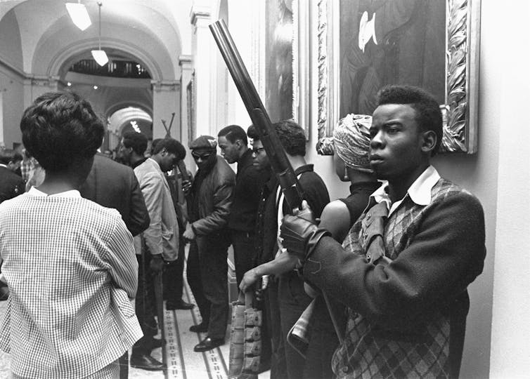 Black and white photo of Black men and women congregating, with some men holding guns.