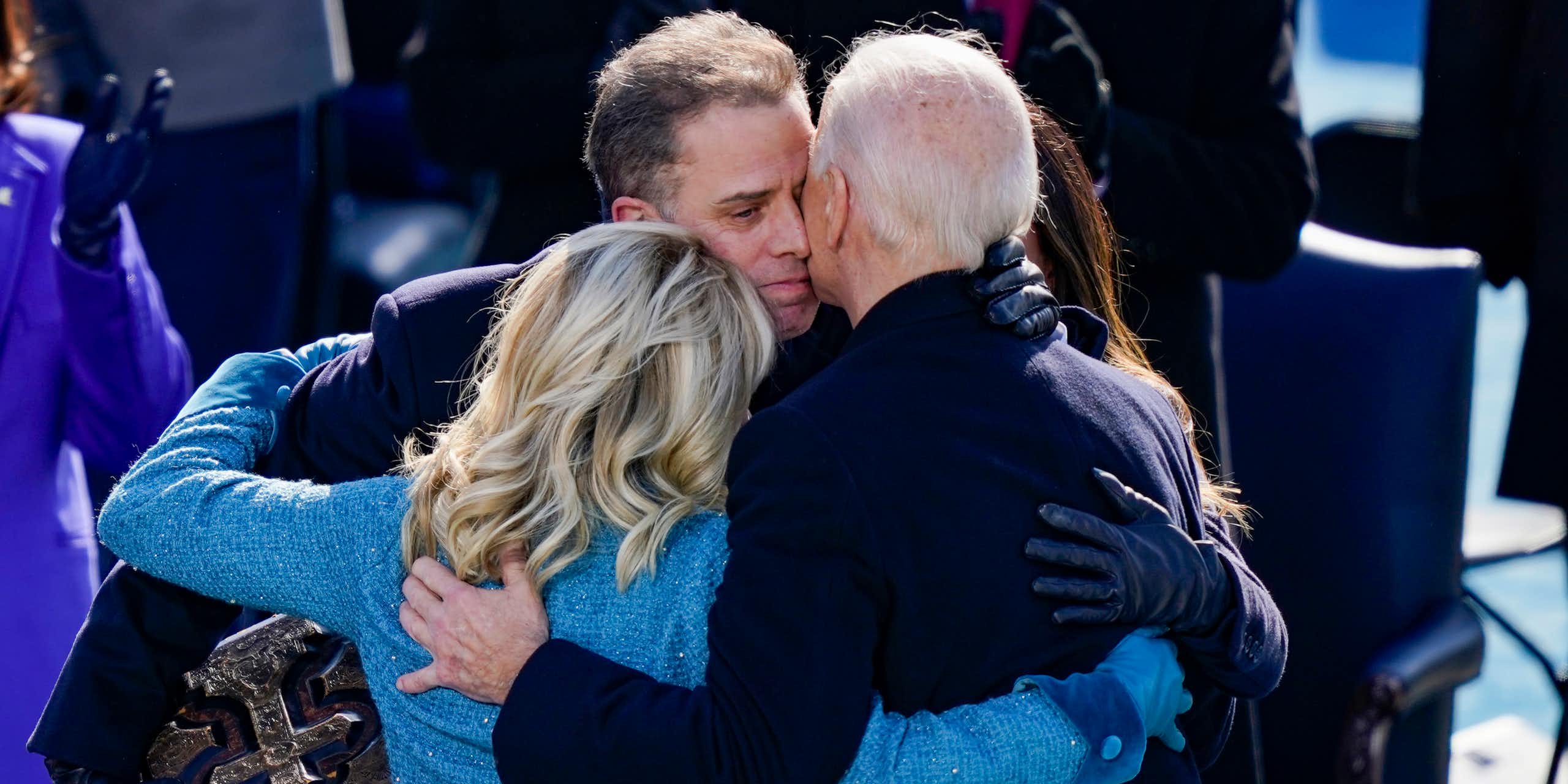 A white man wearing a blue jacket embraces another man with white hair and a woman with blond hair. 