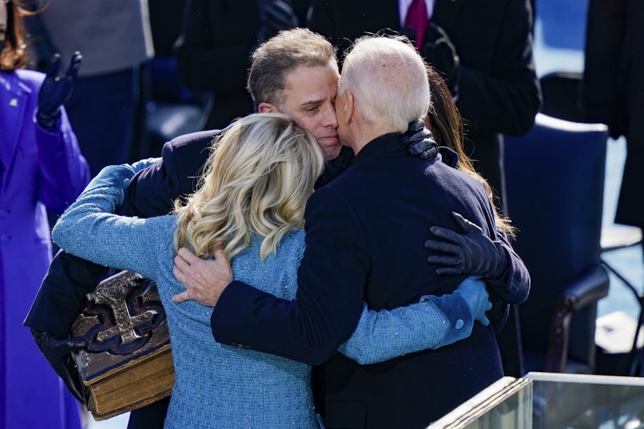 A white man wearing a blue jacket embraces another man with white hair and a woman with blond hair. 