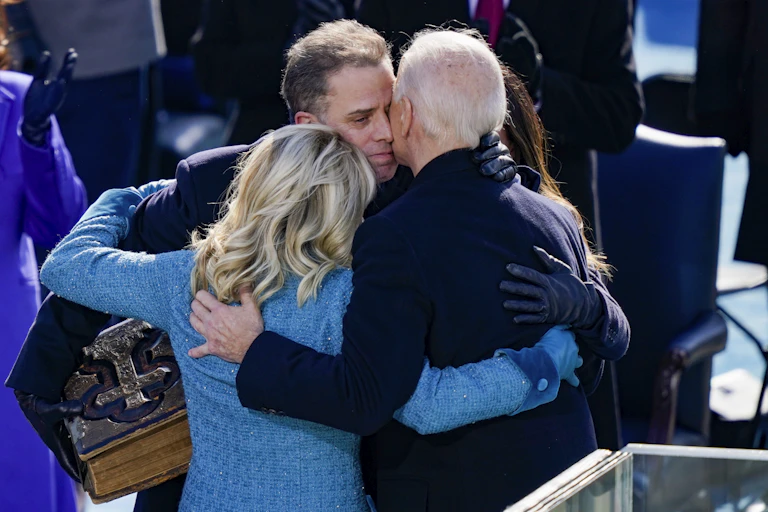 A white man wearing a blue jacket embraces another man with white hair and a woman with blond hair.