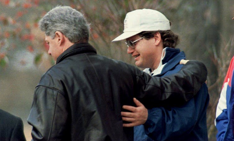 The side profiles of two men are seen as they have their arms around each other - one is Bill Clinton and the other is a man wearing a white hat.