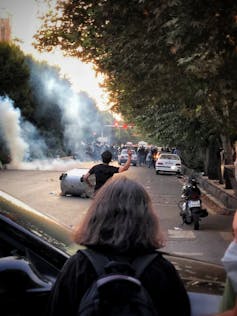 A protestor throwing something at police with a woman in the foreground with her hair free.