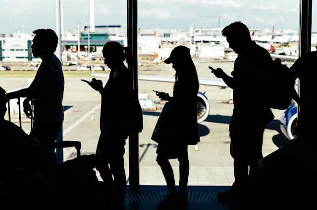 Several people using smartphones silhouetted inside an airport waiting to board a plane visible outside