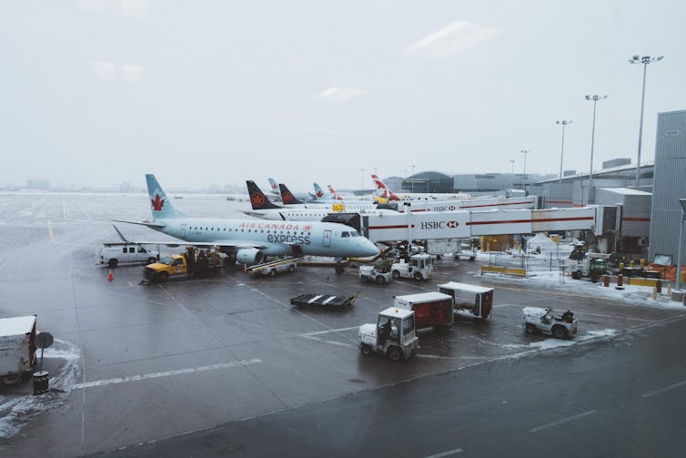 Long view photo of a snowy grey tarmac with an air canada plane and several fuel and other support trucks around it