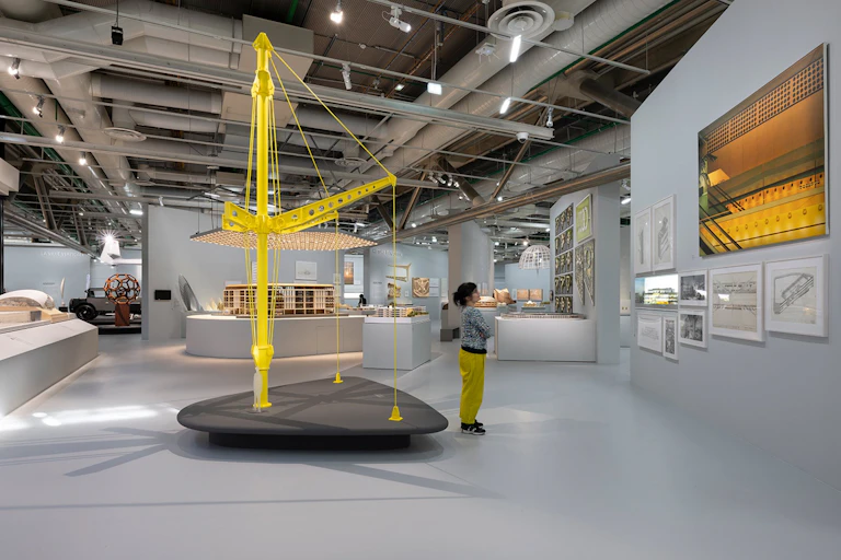 Una mujer observa una pared con planos enmarcados rodeada de objetos y maquetas arquitectónicas.