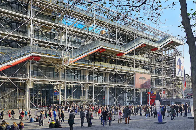 Una plaza llena de gente con un edificio de colores al fondo que deja al descubierto la estructura arquitectónica, las tuberías, las vigas, etc.