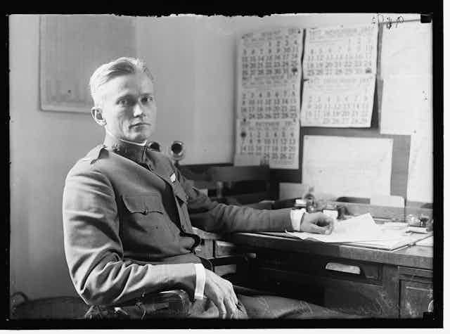 Un hombre rubio de uniforme mira a cámara sentado delante de un escritorio lleno de papeles.