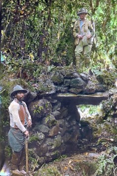 Un hombre vestido de explorador en lo alto de un puente mientras otro hombre mira a cámara desde el camino de debajo.