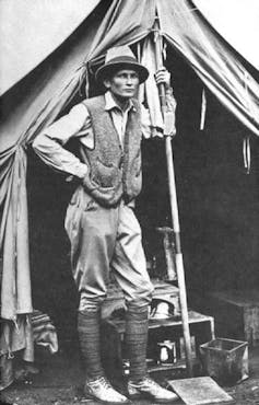 Photographie en noir et blanc d’un homme habillé en explorateur s’appuyant sur un poteau dans sa tente